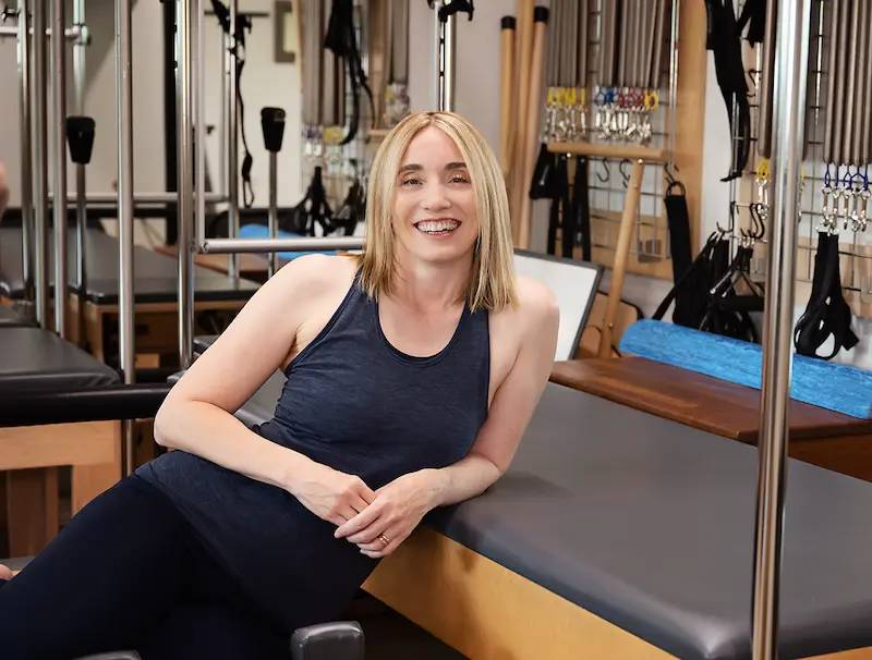 A woman with shoulder-long blonde hair and a black tank top in her physical therapy studio. She is leaning against a black table.