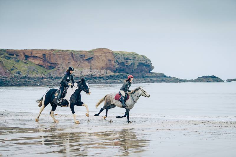 Two young girls ride horses down the beach in Berwickshire
