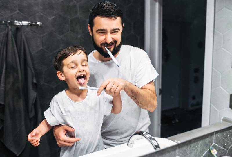 Father and toddler boy brush their teeth in dark bathroom