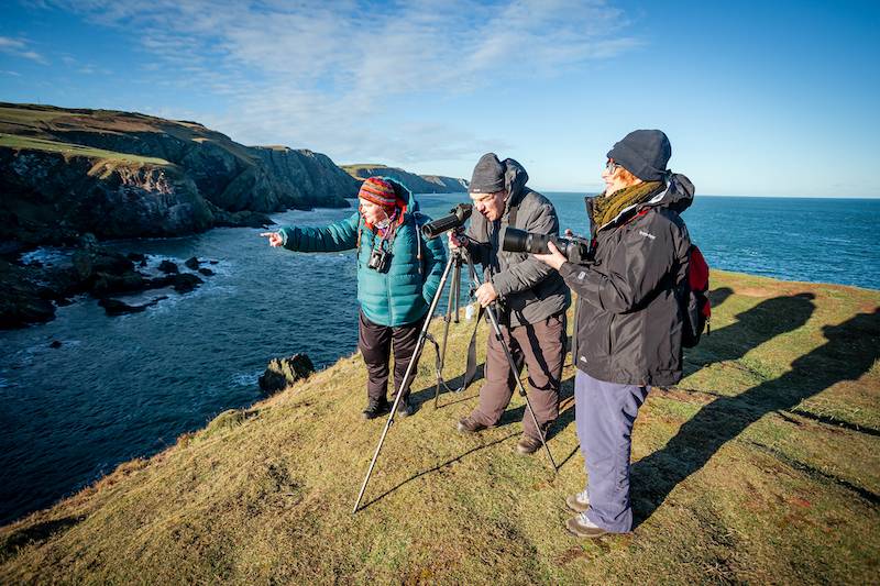 Three people stand on a cliff observing nature through long range cameras with sea in background