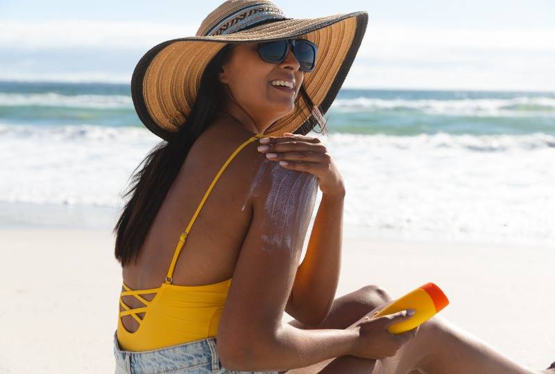 Woman in sunhat and swimsuit on beach puts on sun cream
