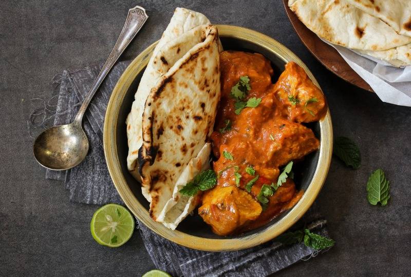 Bowl of butter chicken and naan bread sitting next to a spoon and halved limes