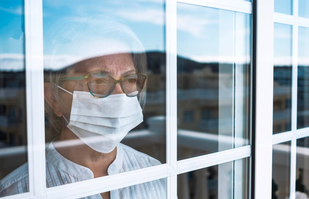 An older woman wearing a face mask staring out of a window during lockdown