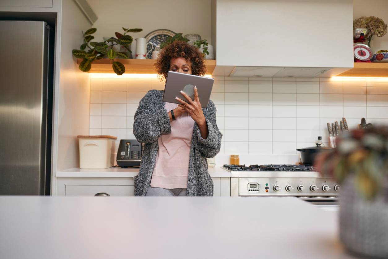 An older brown skinned woman with curly hair using a tablet in the kitchen