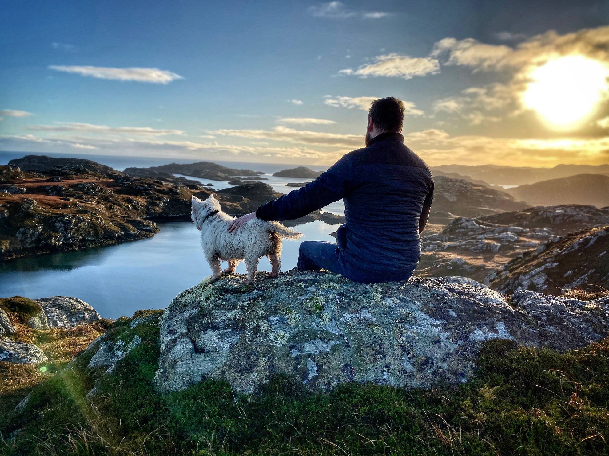 A man with his back to the camera sat on a rock in the Outer Hedrides, while petting his dog